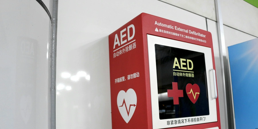 A red AED (Automated External Defibrillator) cabinet mounted on a white wall, featuring a screen with 'AED' text, a heart symbol with an ECG line, and instructions in Chinese and English, including a warning about unauthorized use. The cabinet is equipped with a handle and emergency contact information.