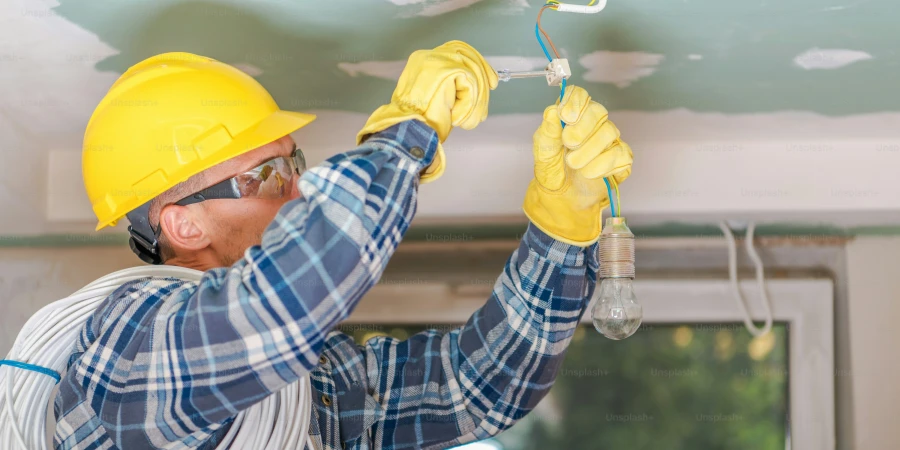 An electrician wearing a yellow hard hat and gloves installs a light bulb into a ceiling fixture, surrounded by exposed wiring in a room under construction.