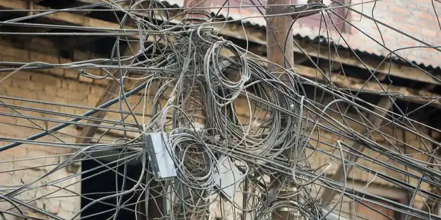 A chaotic mass of tangled electrical wires and cables hanging from a wooden pole and attached to a brick building with a corrugated metal roof, including a small electrical box amidst the clutter.