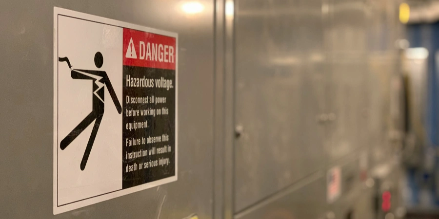 A warning sign with a red and white label displaying a black silhouette of a person being electrocuted, with the text "DANGER Hazardous voltage. Disconnect all power before working on this equipment. Failure to observe this instruction will result in death or serious injury," affixed to a metallic industrial panel.