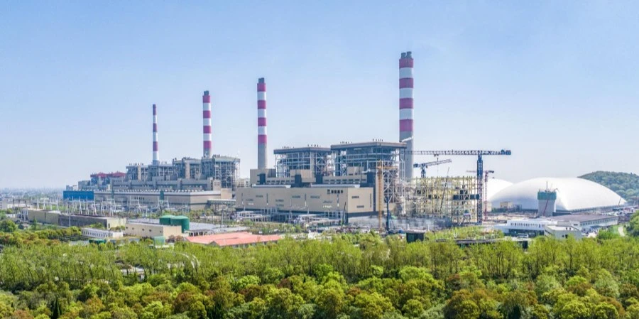 A large coal power plant with multiple chimneys featuring red and white stripes, surrounded by lush green trees and a construction crane, with a clear blue sky above and a white dome structure in the background.