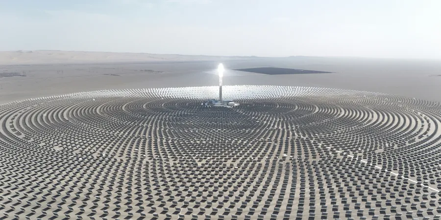 An aerial view of a concentrated solar power plant with a central tower emitting a bright light, surrounded by a vast array of mirrors arranged in concentric circles in a desert landscape.