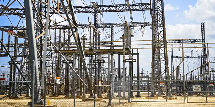 A complex high-voltage electrical substation with numerous metal towers, wires, and insulators, enclosed by a chain-link fence, under a partly cloudy sky.
