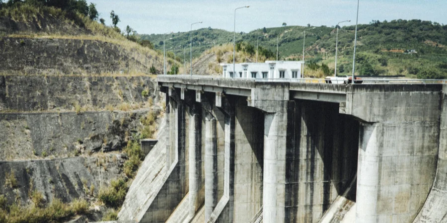 A large concrete hydroelectric power plant with multiple spillways, situated in a hilly landscape with terraced hills and greenery, featuring a road and small buildings on top.