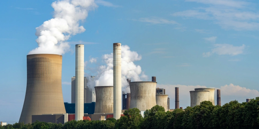 A large industrial coal power plant with multiple cooling towers and smokestacks emitting white smoke, set against a clear blue sky, with green trees in the foreground.