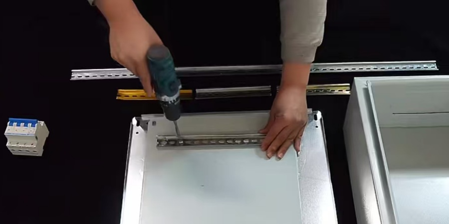 A person using a power drill to install a slotted metal strip onto a panel, with electrical components and a metal enclosure nearby.