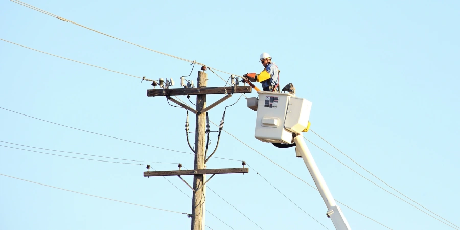 A lineman in a safety helmet and work gear operating from a cherry picker, repairing or maintaining electrical wires on a wooden utility pole under a clear blue sky.
