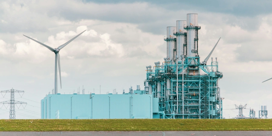 A landscape featuring a modern industrial power plant with multiple chimneys and a blue structure, accompanied by wind turbines and power lines under a cloudy sky.