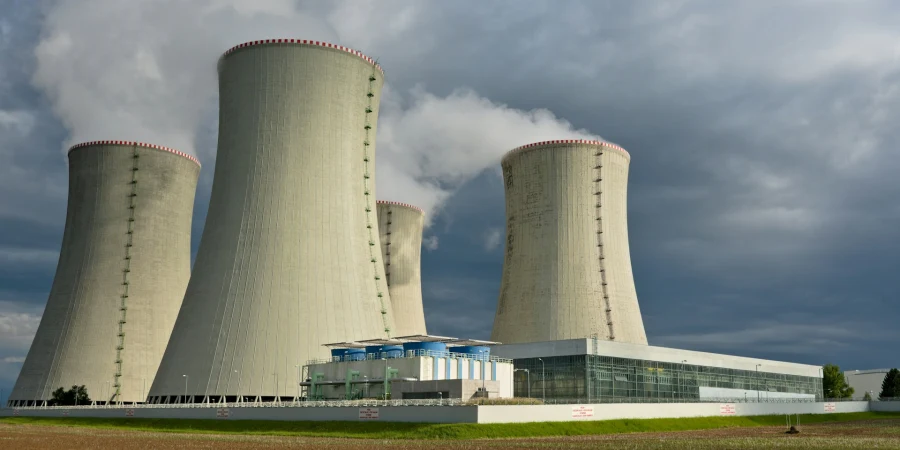 A nuclear power plant featuring three large cooling towers with red and white tops, emitting steam under a cloudy sky, with a modern building and green field in the foreground.