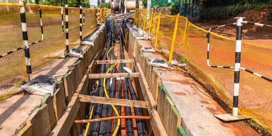 A deep trench lined with wooden supports, containing multiple colorful underground cables (yellow, black, and orange), surrounded by safety netting and yellow barriers with black stripes, set against a backdrop of greenery and a construction site.