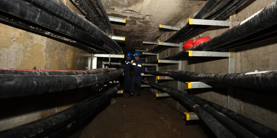 Two workers wearing blue uniforms and helmets inspecting a narrow underground tunnel lined with multiple black cables on metal racks, with red fire extinguishers placed along the shelves, and a rough concrete ceiling.