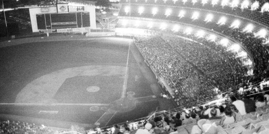 A black and white photo of Shea Stadium in darkness during the bottom of the sixth inning of a game against the Chicago Cubs, July 13, 1977, due to a citywide power failure in New York City, with the field and stands dimly visible.