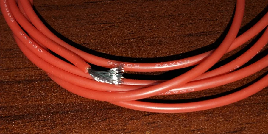 A close-up of several coiled red insulated 20 AWG copper wires on a wooden surface, with one wire showing a damaged section where the insulation is stripped, exposing the silver-colored copper strands inside. The wires are labeled with