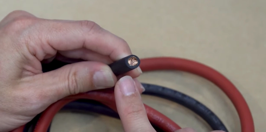 A hand holding a section of 4 AWG copper wire with black insulation, showing the exposed copper end. In the background, coiled red and black insulated wires rest on a beige surface.