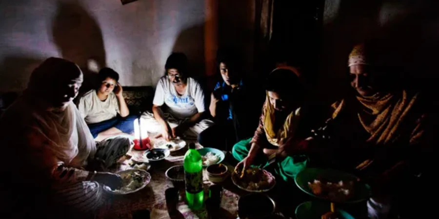 A photo of a Kashmiri Muslim family eating dinner together after breaking their Ramadan fast during a blackout in Srinagar, India, on August 1, 2012, with a dimly lit room, a lantern providing light, and plates of food on the floor.