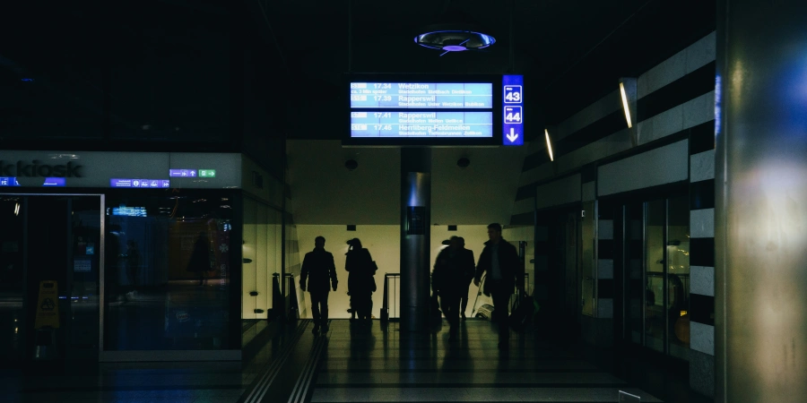 A dimly lit airport terminal during a blackout, showing silhouettes of people walking with luggage, a digital departure board with limited visibility, and a dark, empty hallway with signs and a kiosk in the background.