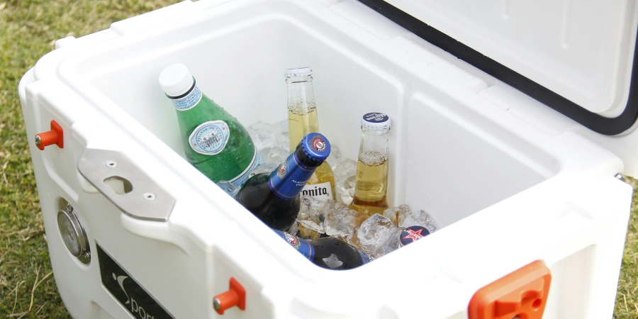 An open white cooler filled with ice, containing various bottled drinks including sparkling water, beer, and soda, placed on a grassy surface.