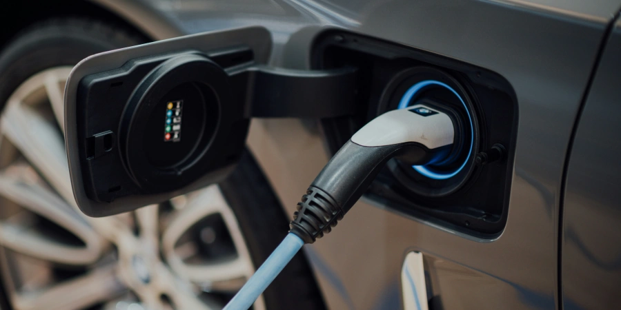 A close-up of an electric vehicle charging port on a gray car, with a charging cable plugged in. The port has a blue illuminated ring and a small display with colored indicators. The car's wheel and part of the body are visible in the background.
