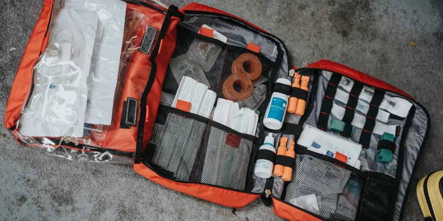 An open orange emergency medical kit on the ground, filled with various supplies including bandages, syringes, vials, and tourniquets, organized in multiple compartments.