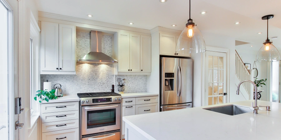 A modern kitchen featuring white cabinets, a stainless steel range and refrigerator, a white countertop with a sink, a range hood with mosaic tile backsplash, pendant lights, and a glimpse of a staircase and greenery, creating a bright and spacious atmosphere.