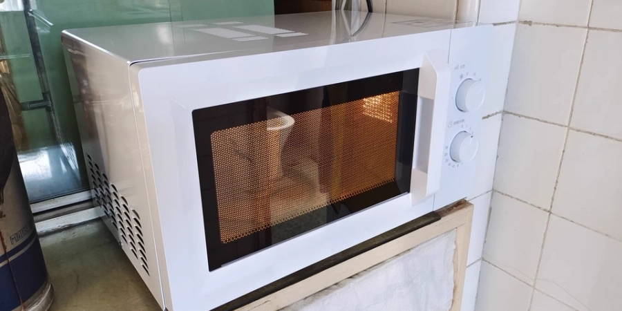 A white microwave oven placed on a countertop against a tiled wall. The microwave has a visible interior with a metal mesh and a cup inside, along with two control knobs on the right side. The surrounding area includes a green wall and a partially visible kitchen setup.