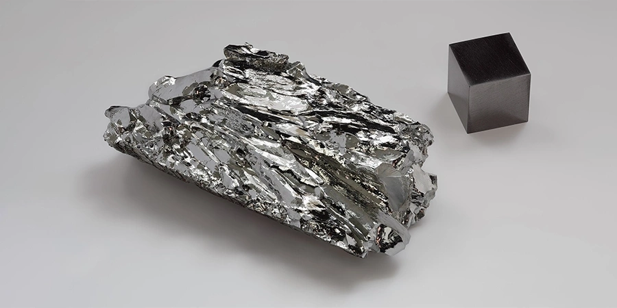 A rough, shiny molybdenum crystal with a metallic, jagged structure, placed next to a smooth, dark molybdenum cube on a plain background.