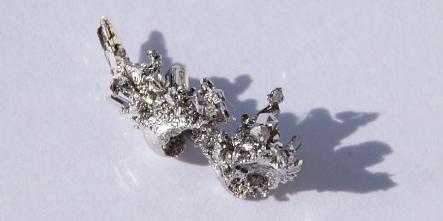 A small, intricate palladium crystal with a shiny, metallic surface and jagged structure, casting shadows on a plain white background.