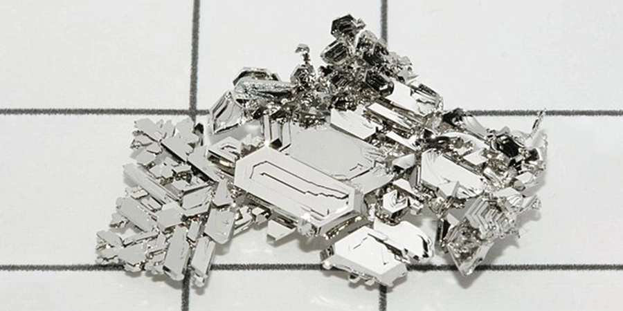 A cluster of shiny platinum crystals with a metallic, jagged structure, displayed on a grid-patterned white background.