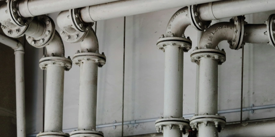 A network of white industrial pipes with flanged joints, arranged vertically and horizontally, mounted against a plain wall in an industrial setting.