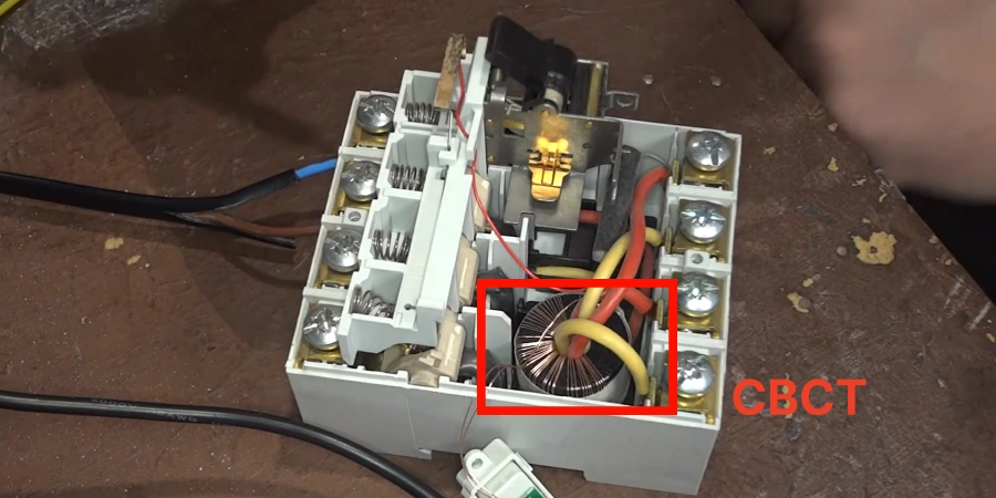 An internal view of a Residual Current Circuit Breaker (RCCB) with a highlighted CBCT (Current Transformer) component in red, surrounded by yellow and red wires, featuring terminals and wiring on a wooden surface, used for detecting leakage currents in electrical systems.
