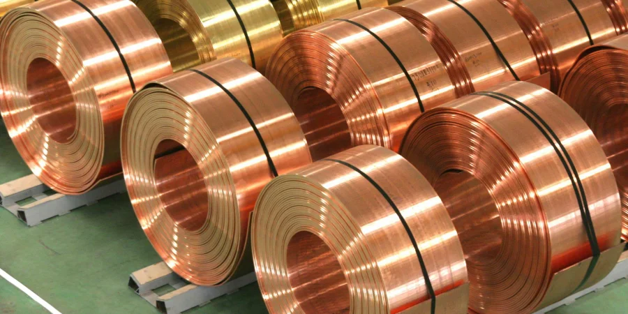 A row of shiny copper coils, some with a golden hue, neatly stacked and secured with black straps on pallets in an industrial storage area.