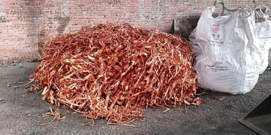 A large pile of reddish-brown scrap copper pieces on a concrete surface, with some white bags labeled in Chinese placed beside it. The background features a brick wall, indicating an industrial or recycling area.