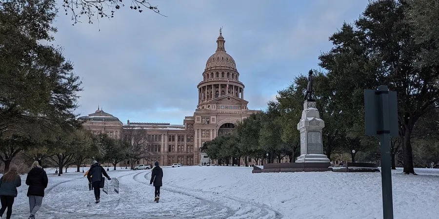 A photo of the Texas Capitol building covered in snow on February 15, 2021, during the 2021 Texas Power Crisis, with people walking on a snow-covered ground, trees, and a statue in the foreground under a cloudy sky.