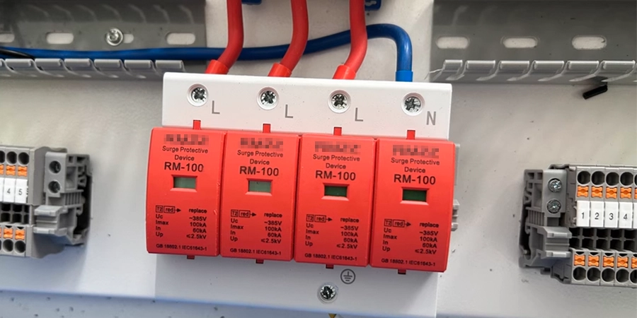 A panel with three red RM-100 surge protective devices mounted on a DIN rail, connected with red and blue wires, displaying green status indicators, used to safeguard electrical systems.