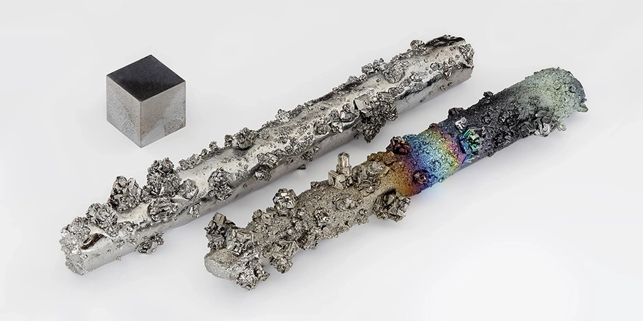 A small, dark tungsten cube alongside two rough tungsten rods, one with a metallic surface and the other displaying colorful oxidation patterns, set against a plain background.