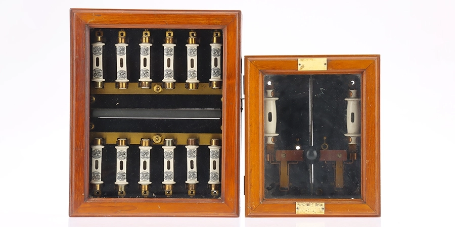 A photograph displaying two vintage wooden fuse box panels from the 20th century. The left panel is open, revealing two rows of ten cylindrical white fuses with metallic ends, mounted on a black and gold interior. The right panel is closed, showing a glass front with two fuses visible inside, along with a central switch or indicator, and a small brass plaque. Both panels have a polished wooden frame, reflecting their historical use in early electrical systems.