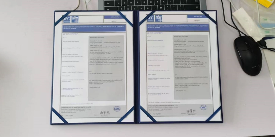 Two CB test certificates for electrical equipment displayed open on a desk, featuring blue covers with the IEC logo and detailed text in multiple sections. A keyboard, mouse, and yellow object are visible in the background, with a laptop partially shown.