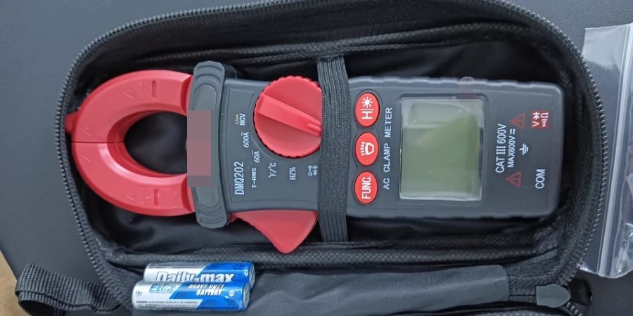 A black clamp meter with a red jaw and dial, featuring a digital display and multiple buttons, stored in an open black protective case. Two blue batteries and a small plastic bag are placed beside it on a dark surface.
