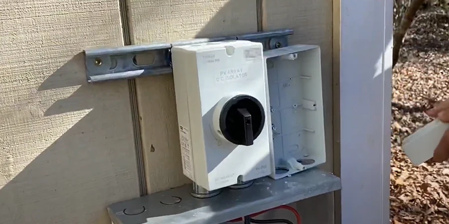A close-up image of a DC isolator mounted on the exterior wall of a building, featuring a white enclosure with a black rotary switch. The isolator is labeled 