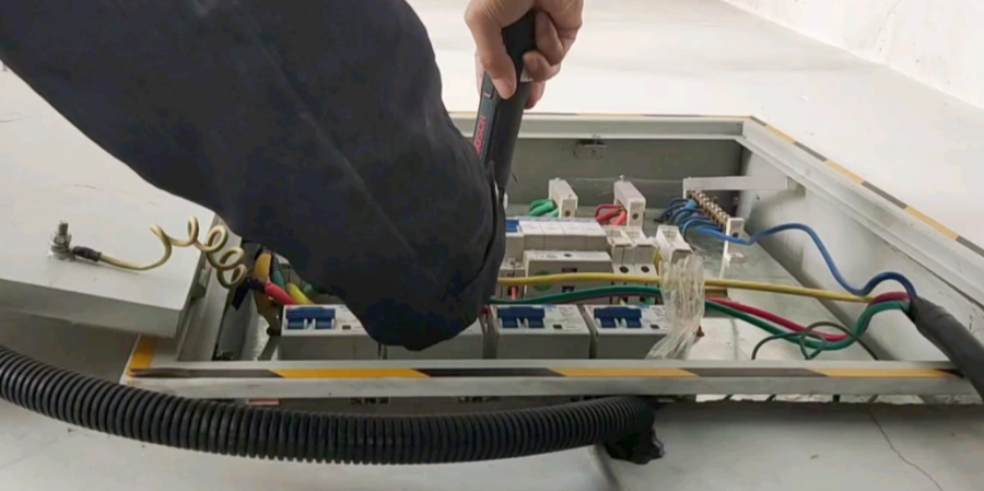 A technician using a screwdriver to replace or inspect a circuit breaker inside an open electrical distribution box. Various colored wires are connected to the terminals, indicating active electrical work.