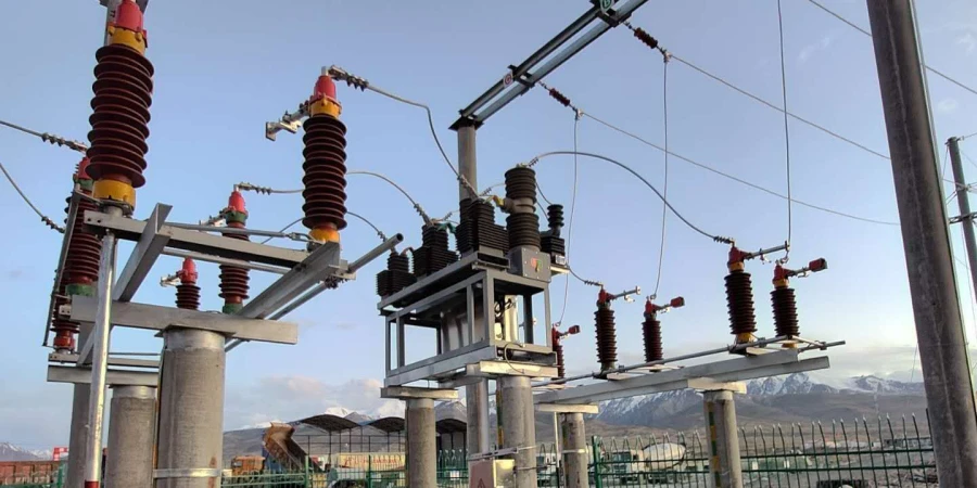 An image of a high-voltage circuit isolator installed in an outdoor substation, featuring multiple red and yellow insulators mounted on a metal framework. The setup includes several circuit breakers and connected wires, supported by concrete pillars. The background shows a clear sky, distant snow-capped mountains, and a fenced industrial area with some equipment and buildings.