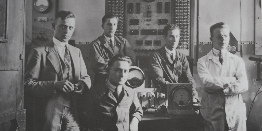 A black-and-white photograph featuring five men in an early 20th-century electrical workshop. The men are dressed in formal suits and work coats, standing and sitting around a table with vintage electrical equipment, including a control panel with gauges and switches on the wall behind them. One man is seated at the table, adjusting a device, while the others stand or lean casually, with one holding a tool. The setting suggests a collaborative environment focused on electrical engineering innovation.