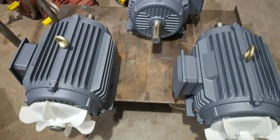 An image featuring three gray industrial electric motors placed on a wooden pallet. Each motor has a ribbed exterior, a white cooling fan at the front, and a gold lifting hook on top. The background shows a workshop floor with some equipment and yellow-handled tools partially visible.