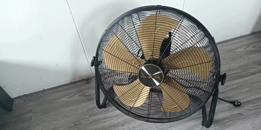 An image of an industrial floor fan with a black metal frame and stand, featuring five golden metal blades encased in a protective wire guard. The fan has a black and gold central hub with a brand label, and a power cord with a plug visible on the floor. The fan is positioned on a wooden floor against a white wall in an indoor setting.