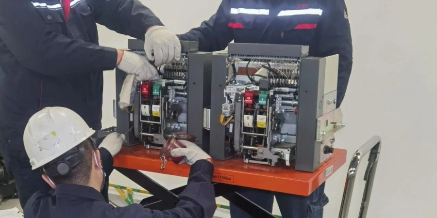 Three technicians in navy blue uniforms and white gloves working on an air circuit breaker mounted on an orange platform. One technician, wearing a white hard hat, is focused on the internal components, while the other assists. The breaker features colorful switches and wiring, with a metal cart visible in the background.
