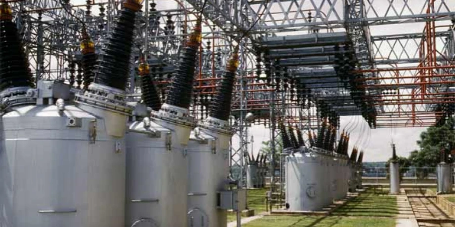 A photograph of an outdoor power substation featuring multiple large oil circuit breakers arranged in a row. The breakers are housed in cylindrical metal tanks with insulators and high-voltage bushings extending from the top. Above the breakers, a complex network of metal frameworks and electrical connections is visible, supporting the high-voltage lines. The scene includes grassy areas and a clear sky, indicating an open, industrial environment designed for electrical power distribution and protection.