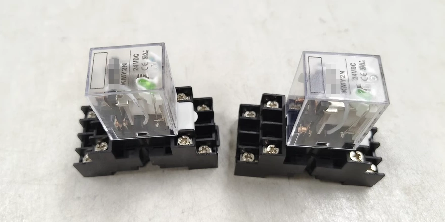 An image showing two 24V DC relays mounted on black bases, placed side by side on a white background. Each relay is encased in a transparent plastic cover, revealing internal components including a green indicator light and metal contacts. The relays are labeled