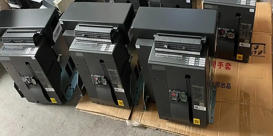 Several black rectangular ACB (Air Circuit Breaker) devices placed on brown cardboard boxes in a warehouse. The devices feature labels, QR codes, small displays with red and green indicators, and ventilation grills, with some Chinese text on the boxes indicating handling instructions.