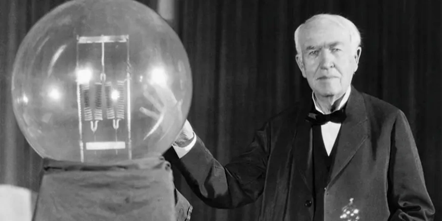 A black-and-white photograph of Thomas Edison, an elderly man with white hair, dressed in a formal suit and bow tie, standing and gesturing toward a large glass light bulb. The bulb, mounted on a pedestal, contains visible filaments glowing inside, showcasing an early incandescent design. The background is dark, focusing attention on Edison and the invention, highlighting his role as a pioneer in electrical innovation.