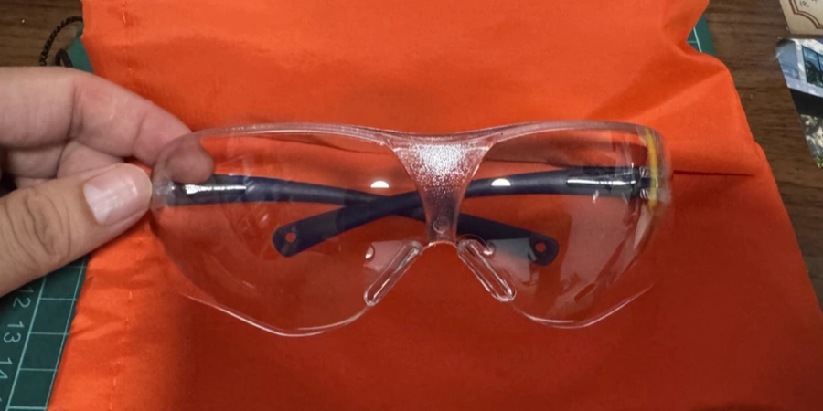 A pair of clear-lens safety glasses with black temple arms, held over an orange cloth. Designed to protect eyes from dust, debris, and impact during industrial or electrical work.